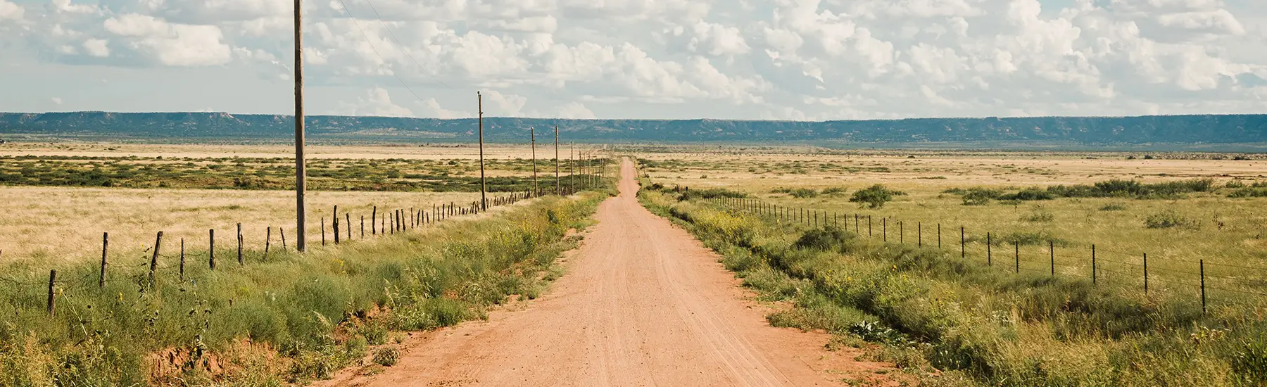 Dirt road lined by wire fences going straight out toward distant hills.